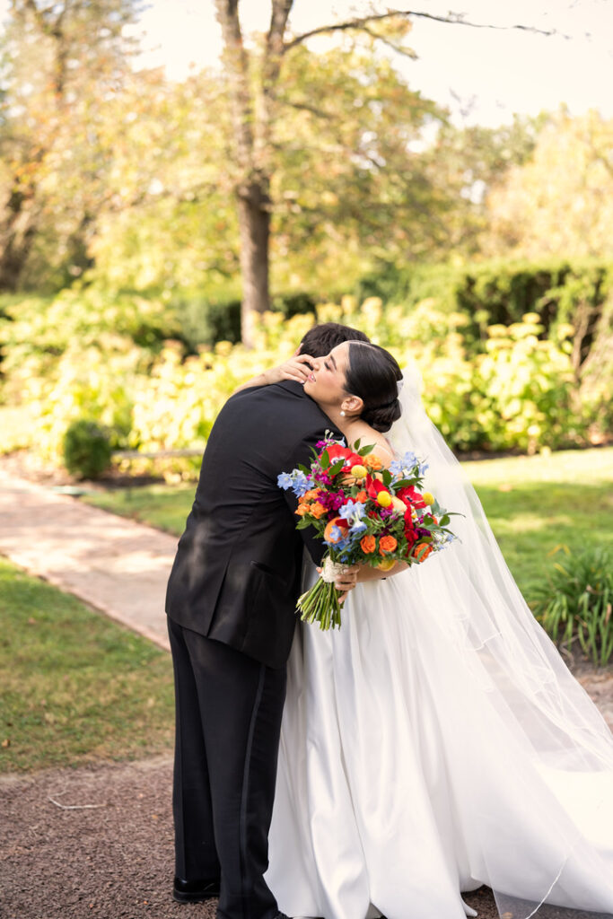 A bride and groom embrace outdoors. The bride holds a colorful bouquet, and both wear formal attire. Trees and greenery are in the background.
