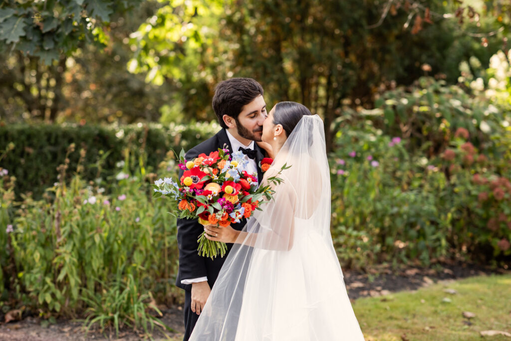 A bride and groom smile at each other outdoors. The bride holds a colorful bouquet, and the groom wears a suit. They are surrounded by lush greenery.