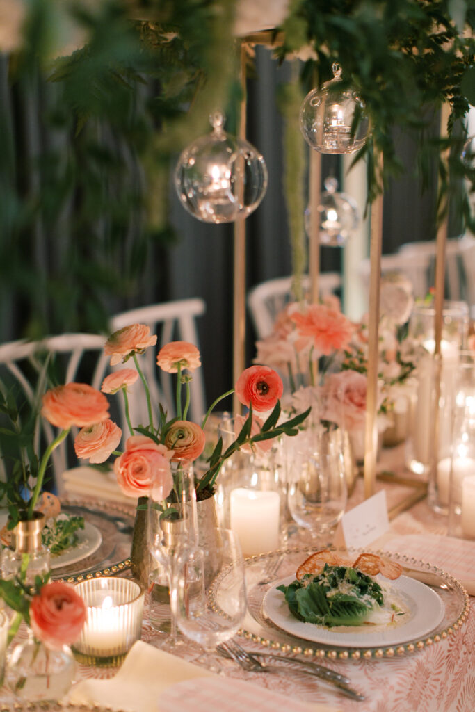 Elegant dining table with floral arrangements, glass ornaments, and candles, reminiscent of a Tillinghouse Wedding. Neatly set plates with salad are surrounded by light pink flowers and greenery, creating a romantic ambiance.