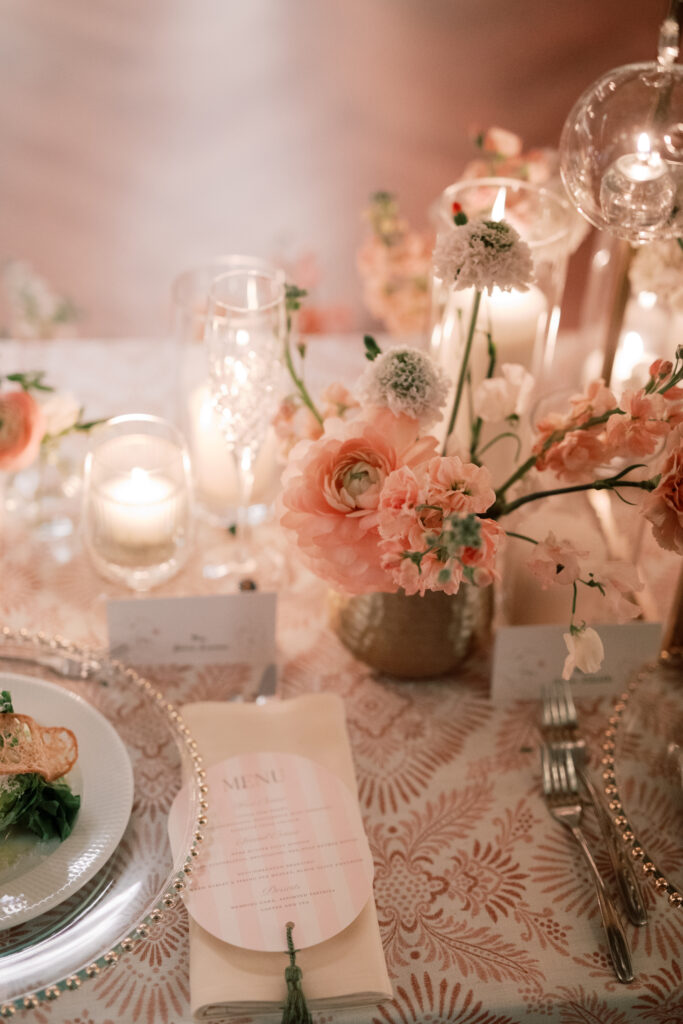 The Tillinghouse Wedding featured an elegant table setting with a pink floral centerpiece, candles, and crystal glassware on a patterned tablecloth. The menu and utensils were neatly arranged alongside, creating an ambiance of romantic sophistication.