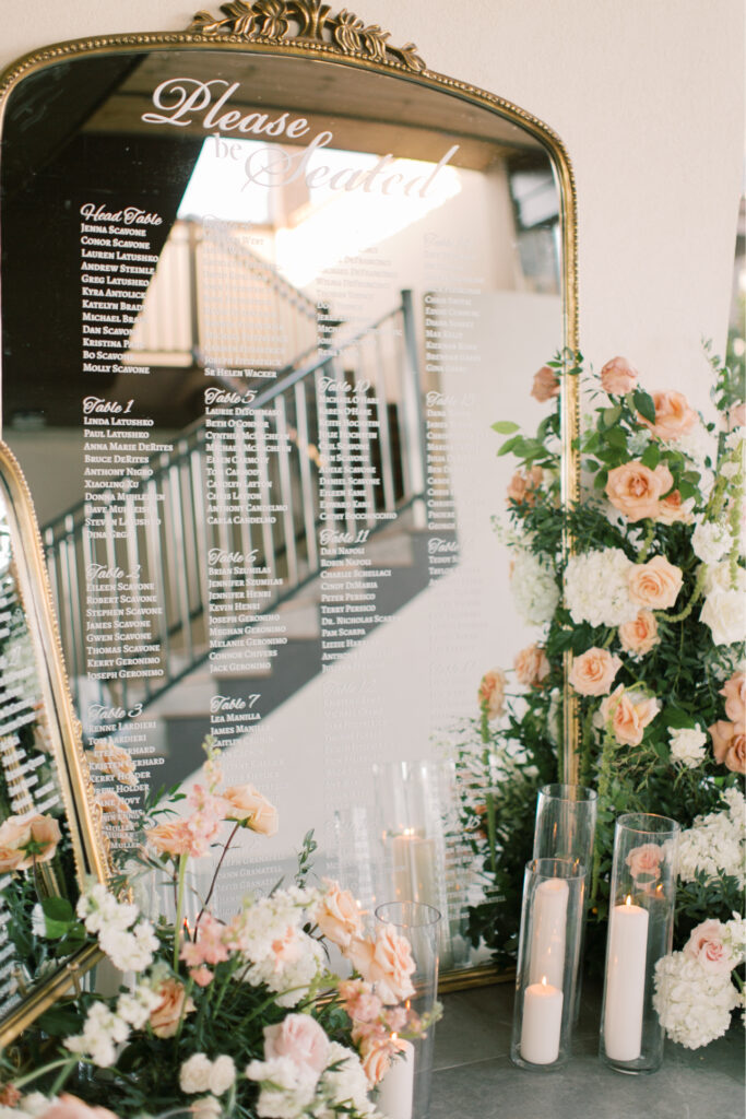 At The Tillinghouse in NJ, a large ornate mirror beautifully displays the guest seating chart, surrounded by pastel flowers and lit candles in glass holders.