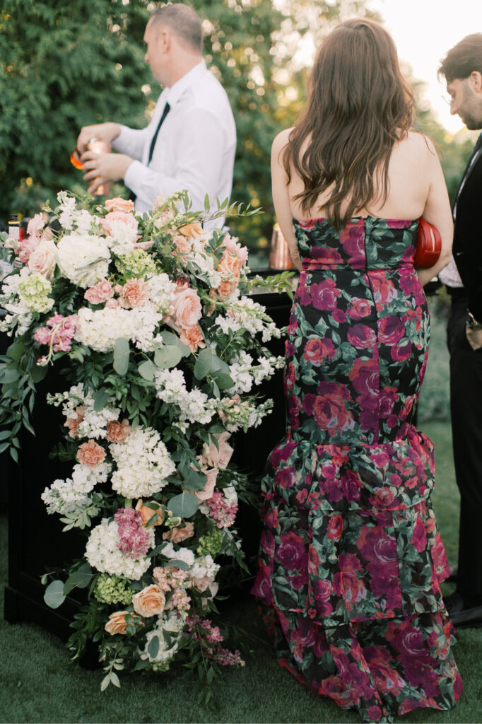 At The Tillinghouse in NJ, a woman in a floral dress stands elegantly by a bar adorned with a grand arrangement of white and pink flowers, while the bartender skillfully prepares drinks in the background.