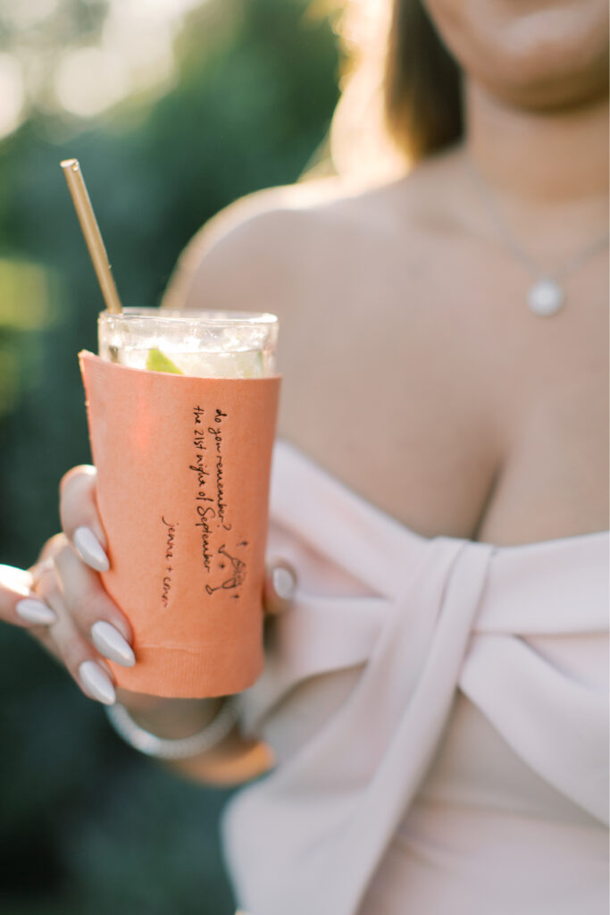 A person at The Tillinghouse in NJ, dressed in a light-colored outfit, holds a drink in a cup with an orange sleeve and straw. A lime wedge perches on the rim, adding a zesty touch to their refreshing beverage.