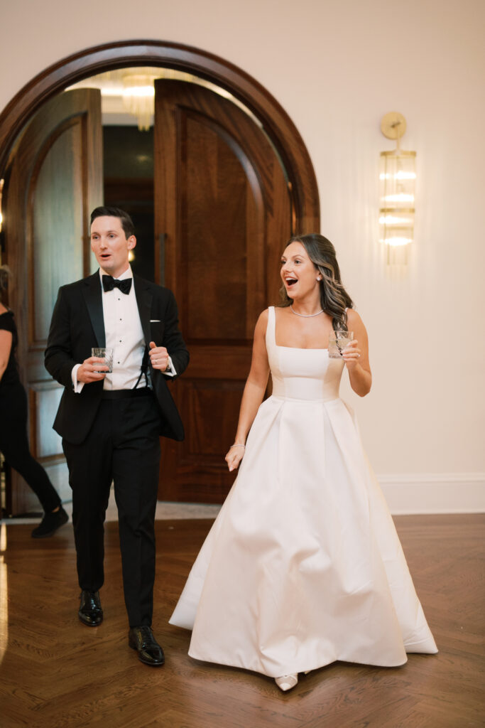 A couple in formal attire, the man in a black tuxedo and the woman in a white gown, walk through a doorway while smiling and holding drinks.