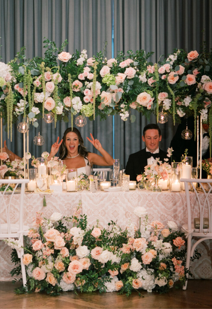 At a beautifully decorated Tillinghouse wedding in NJ, the bride and groom sit at a charming table adorned with flowers and candles. The bride playfully gestures with peace signs, capturing the joyful spirit of their special day.
