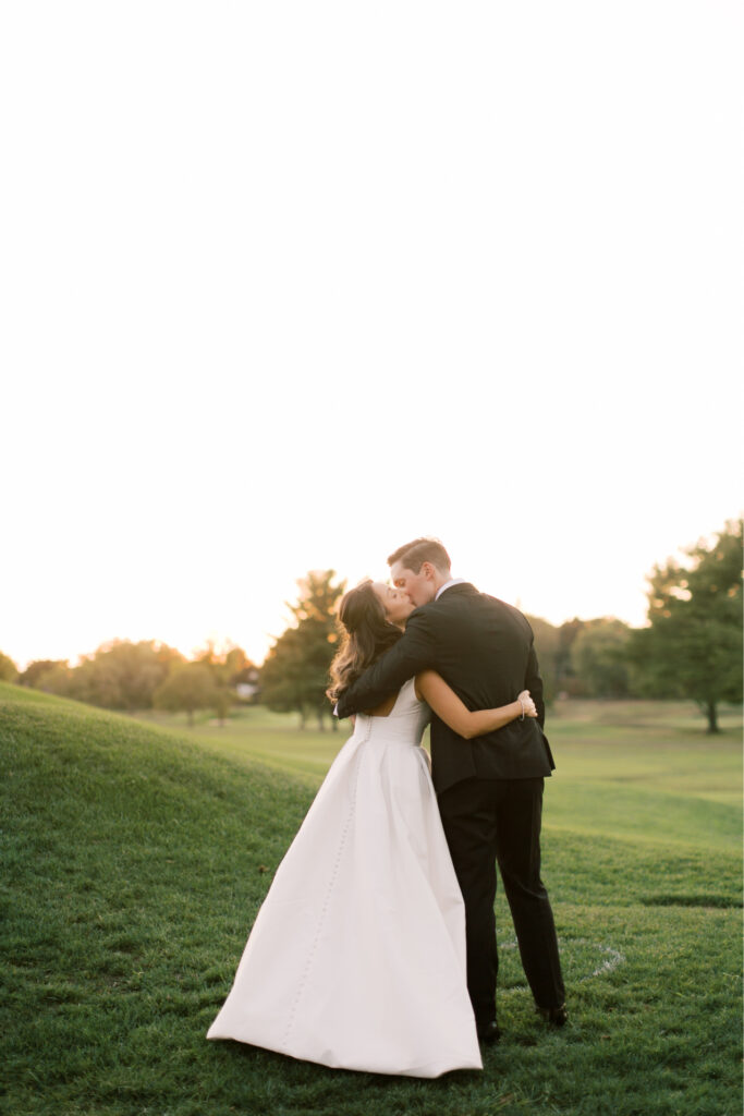 A couple in formal attire embraces and kisses in a grassy field at The Tillinghouse in NJ during sunset, with trees silhouetted beautifully in the background.