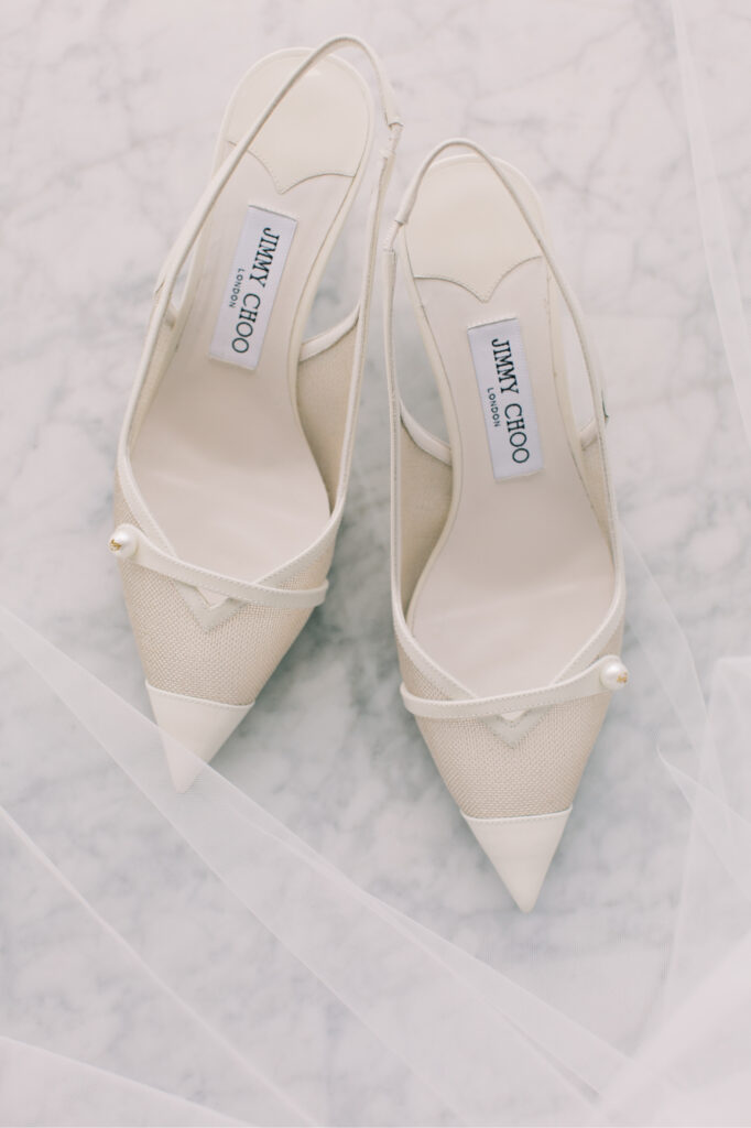 White Jimmy Choo pointed-toe slingback heels with mesh details and pearl accents, placed on a light marble surface with sheer fabric around. Details wedding prep photo.