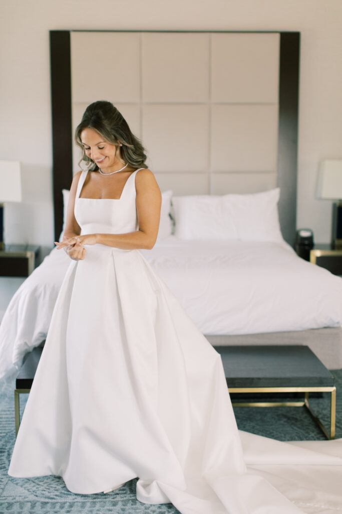 A woman in a white wedding dress stands indoors in front of a bed, looking down and smiling while adjusting her bracelet.