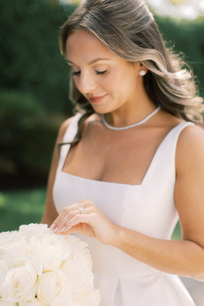 A woman in a white dress holds a bouquet of white roses outdoors, showcasing a ring on her finger. She has a necklace and earrings, with trees visible in the background.