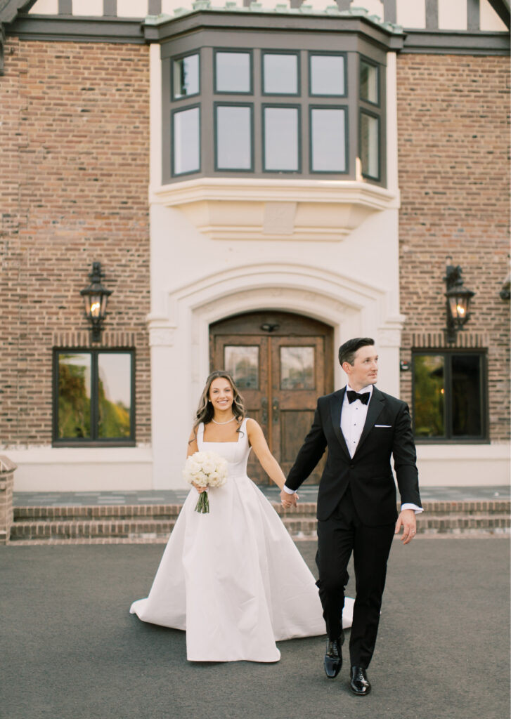 A bride and groom, dressed formally, hold hands and smile while walking in front of a brick building with a decorative window.