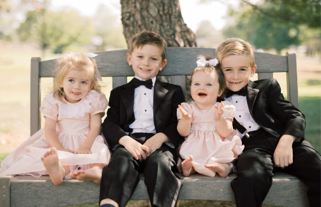 Four children sit on a bench outdoors, with trees in the background. The two boys are in tuxedos, and the two girls are in pink dresses, all smiling at the camera. Flower girls and ring bearers at a wedding.