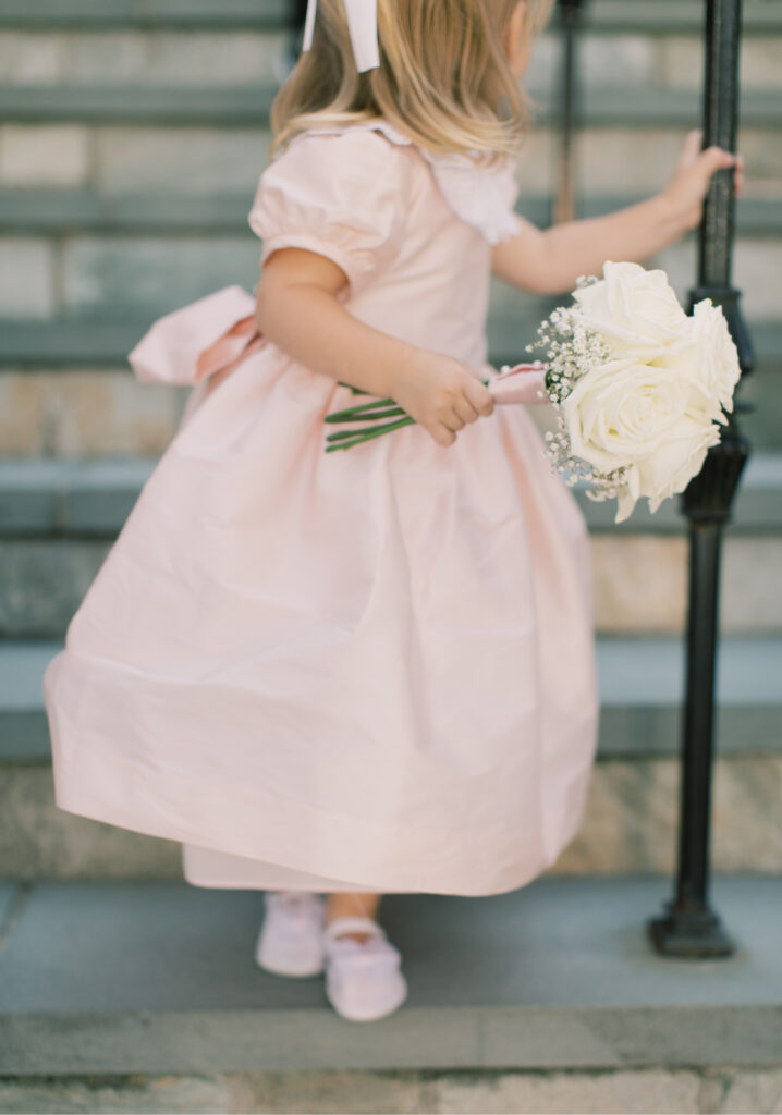 A young girl in a light pink dress holds a bouquet of white roses while standing on stone steps. A flower girl ready for a wedding.