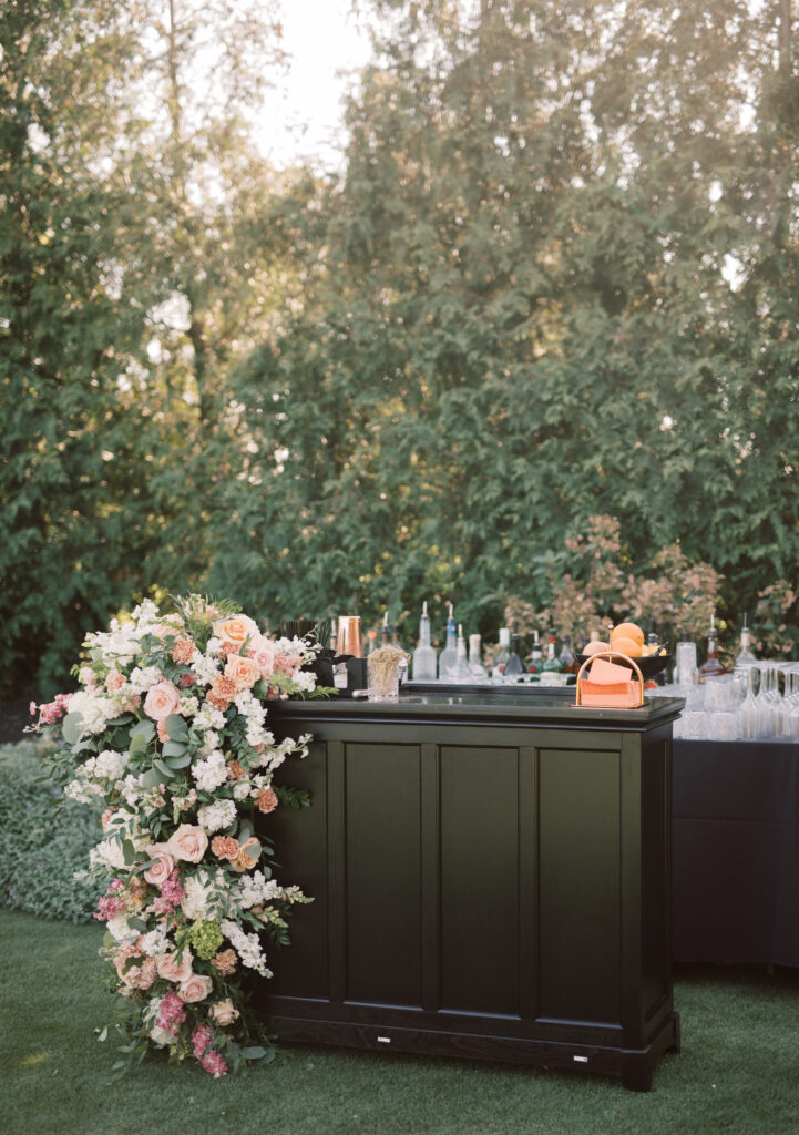 Outdoor bar setup at The Tillinghouse in NJ, featuring a black counter adorned with pink and white flowers on the side. Various bottles and glasses are arranged on top, set against a backdrop of lush green trees.