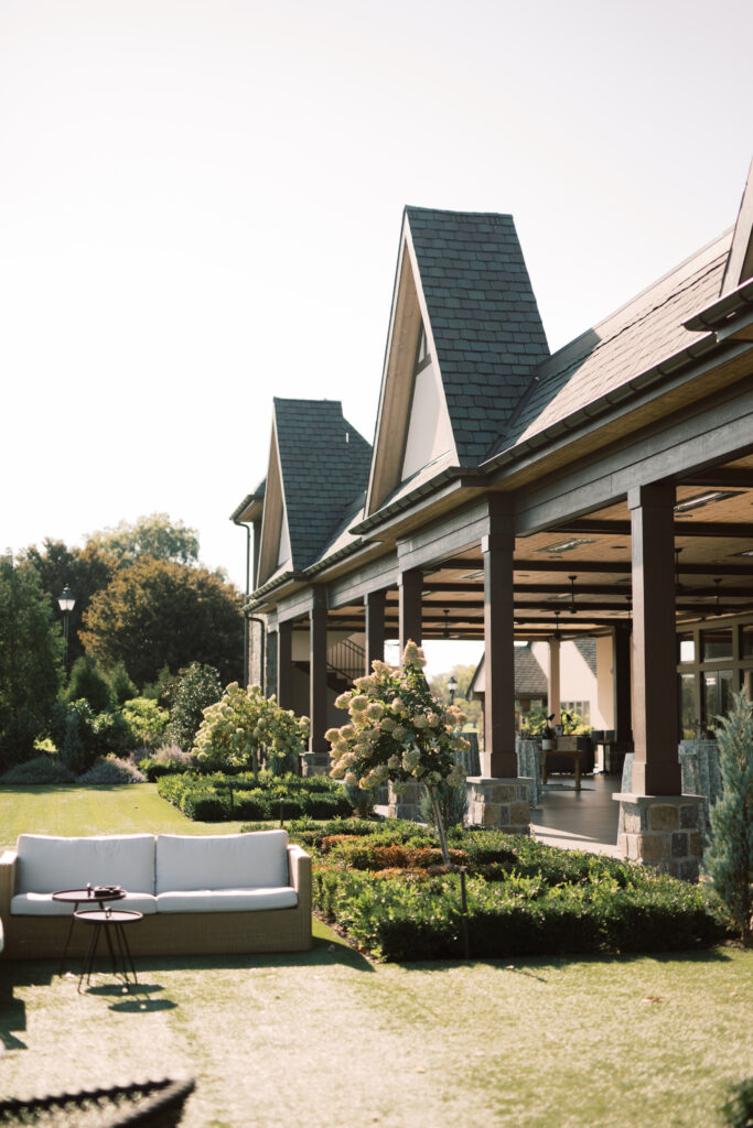 The Tillinghouse in NJ with pointed roofs, a covered patio, and a landscaped lawn featuring bushes and a sofa seating area in the foreground is perfect for an enchanting wedding ceremony.