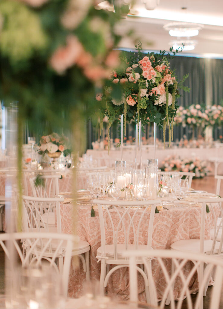 The elegant Tillinghouse Wedding setup featured round tables draped in pink lace tablecloths, complemented by tall floral centerpieces, white chairs, and candle decorations that added a gentle glow to the softly lit room.